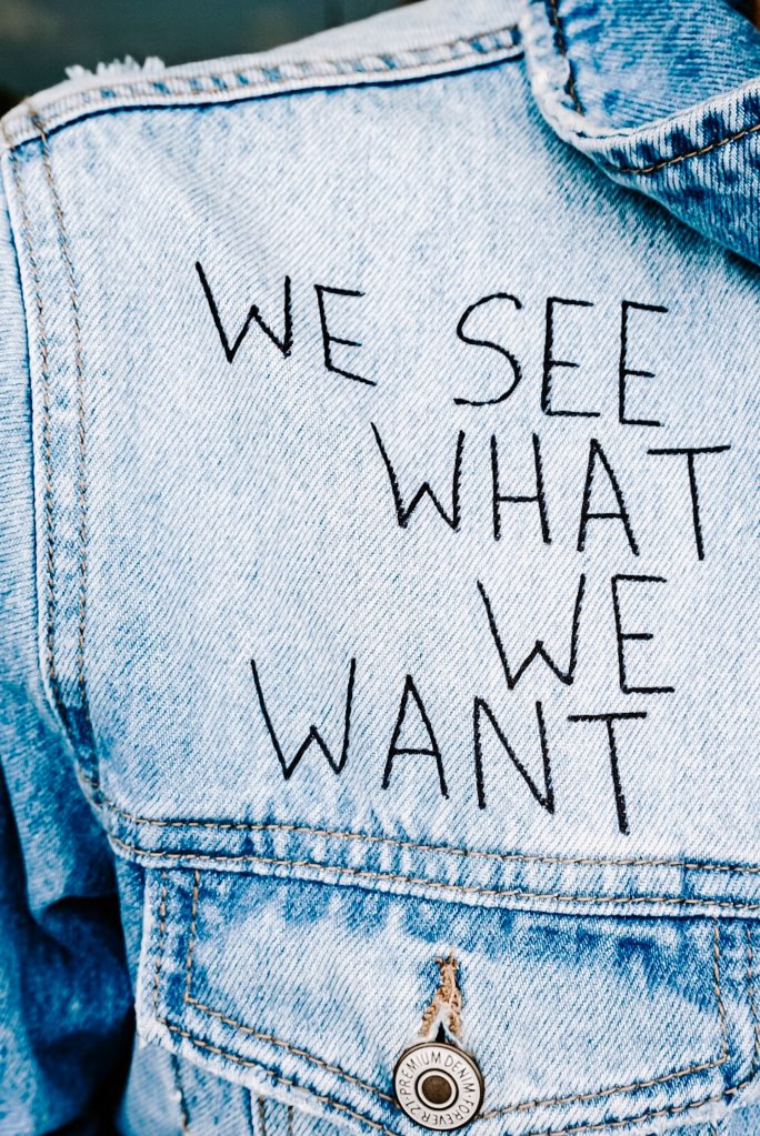 Denim shirt with black lettering that reads, "we see what we want"  Caption: what do you see in the world?